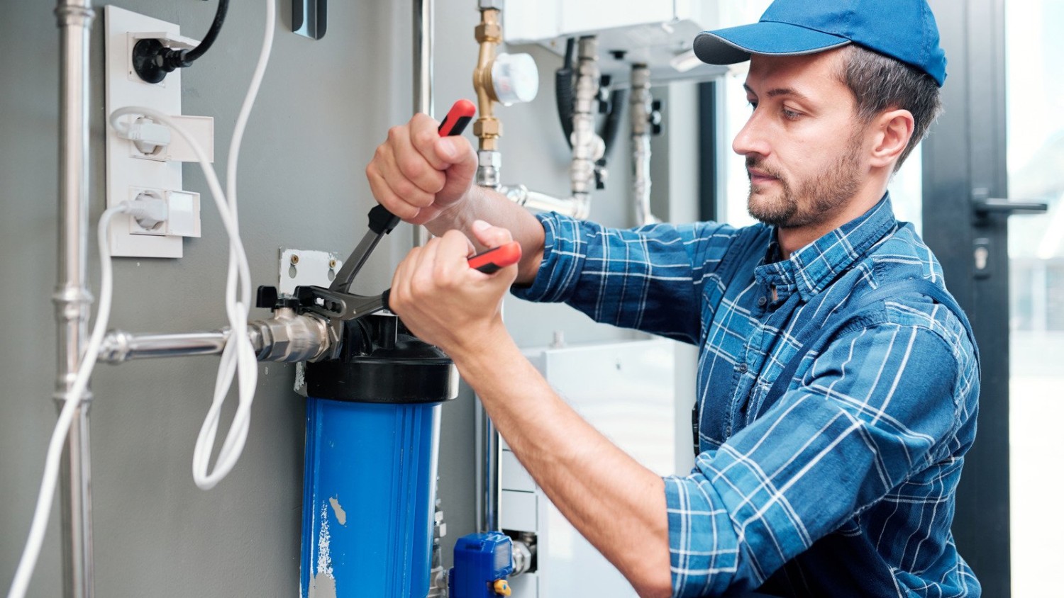 A technician in a plaid blue shirt using a wrench to adjust a blue water filter housing unit connected to a residential water heating system.