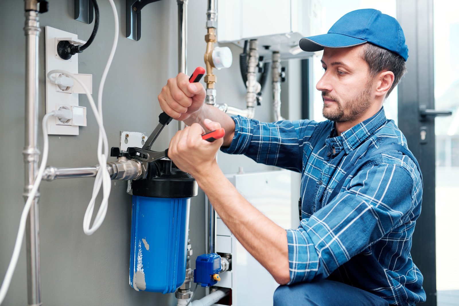 A technician in a plaid blue shirt using a wrench to adjust a blue water filter housing unit connected to a residential water heating system.