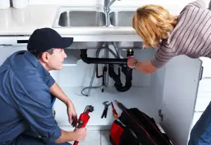 A plumber in a blue uniform and black cap kneels under a double kitchen sink, holding a pipe wrench while a woman points to the black PVC drainage pipes. A tool bag and various wrenches are laid out on the floor of the cabinet.